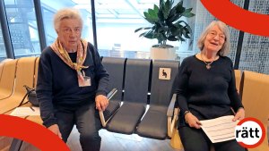 Två kvinnor på en kurvad bänk. Flera säten är bruna, men i mitten finns blå platser med armstöd och en skylt med ikonen för gravida.