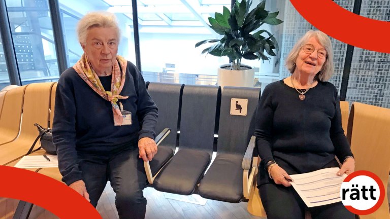 Två kvinnor på en kurvad bänk. Flera säten är bruna, men i mitten finns blå platser med armstöd och en skylt med ikonen för gravida.