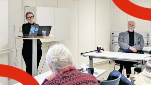 Karin Aronsson står vid ett högt bord, några andra personer i lokalen sitter och lyssnar på henne prata.