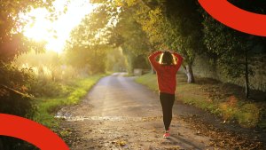 Solen skiner genom träd och buskage, intill en grusväg. På vägen går en kvinna i mjuka träningskläder.