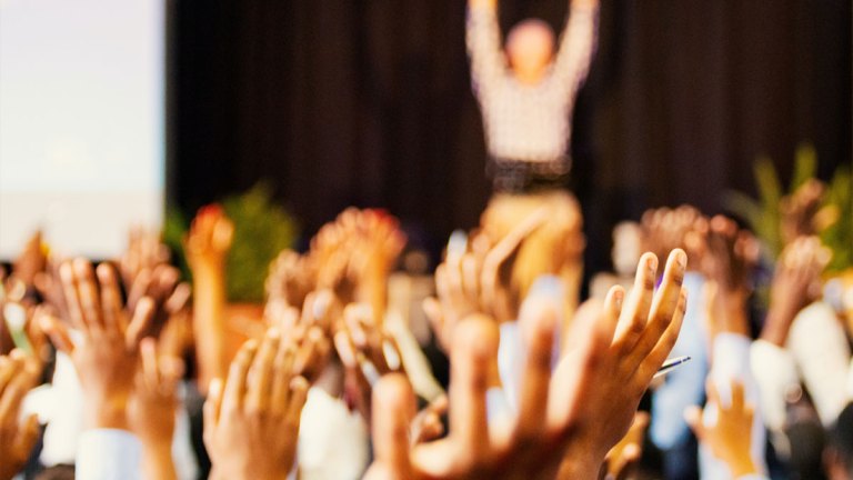 Personer i ett möte håller upp flera uppsträckta händer. I bakgrunden står en person på ett podium.