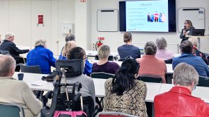 Karin står intill en presentation som visas på en vägg. Hon håller en mikrofon. Framför henne sitter en publik och lyssnar.