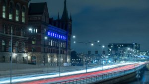 En kvällsbild där bilar flödar förbi på en väg, så snabbt att endast strecken efter lamporna syns. I bakgrunden lyser Nordstedtshusets blå neonbokstäver fram husets namn.