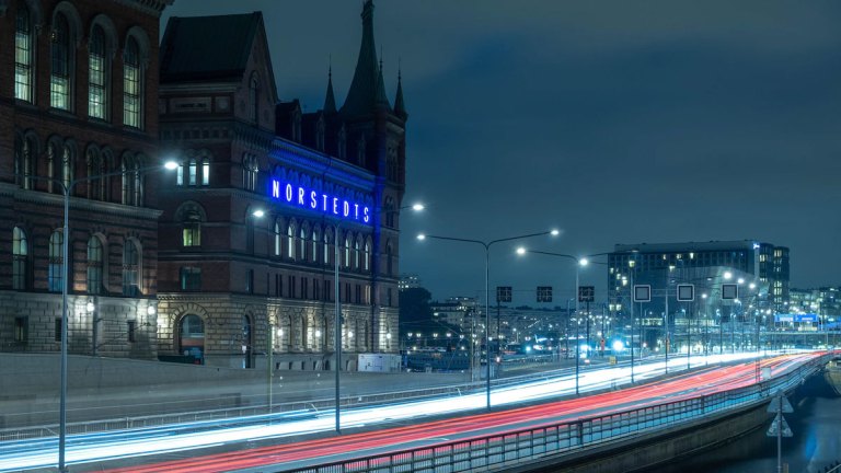 En kvällsbild där bilar flödar förbi på en väg, så snabbt att endast strecken efter lamporna syns. I bakgrunden lyser Nordstedtshusets blå neonbokstäver fram husets namn.