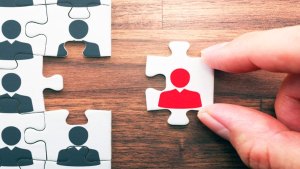 Ett pussel ligger på ett bord. En hand ska sätta sista biten på plats. Motivet på pusslet är figurer av människor.