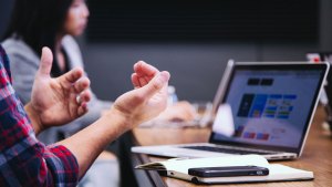 Gestikulerande händer i förgrunden. Resten av personen syns inte, men den har en laptop, anteckningsbok och mobil intill sig. I bakgrunden sitter en annan person och lyssnar.