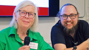 Annika och Tjarles sitter intill varandra och ler glatt. Annika håller en mikrofon.