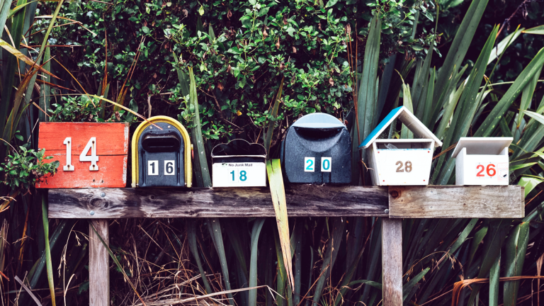 Meddela nya kontaktuppgifter! Flera olika brevlådor är uppsatta på rad på en träram. I bakgrunden rik grönska.