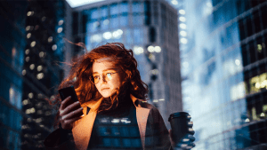 En kvinna med rött, lockigt hår står framför höga skyskrapor. Hon ser på sin mobiltelefon.