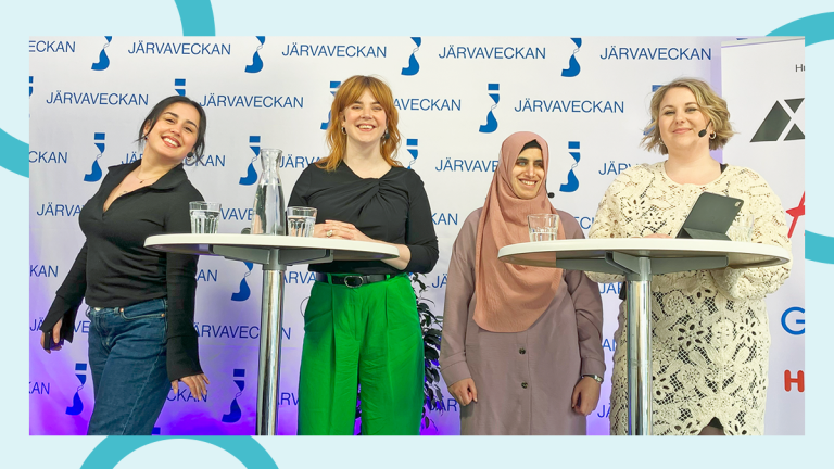 Fyra unga kvinnor står samlade kring två ståbord. De ser glada ut. I bakgrunden hänger en Järvaveckan-back drop.