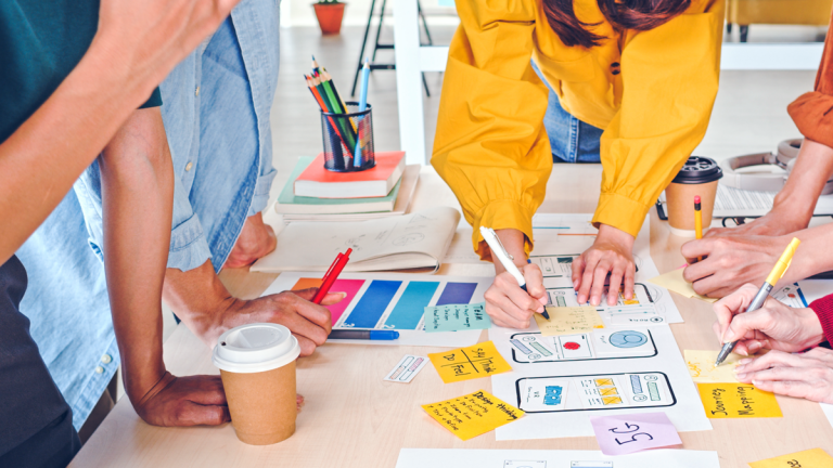 Flera personer står lutade över ett bord med anteckningar, postit-lappar och kaffemuggar.