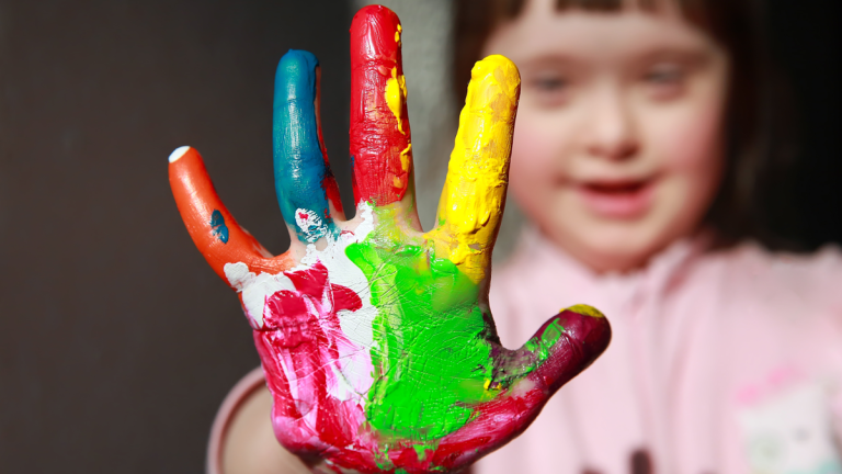 En glad flicka med Downs syndrom stäcker ut sin hand, som är täckt av målarfärg i flera olika nyanser.