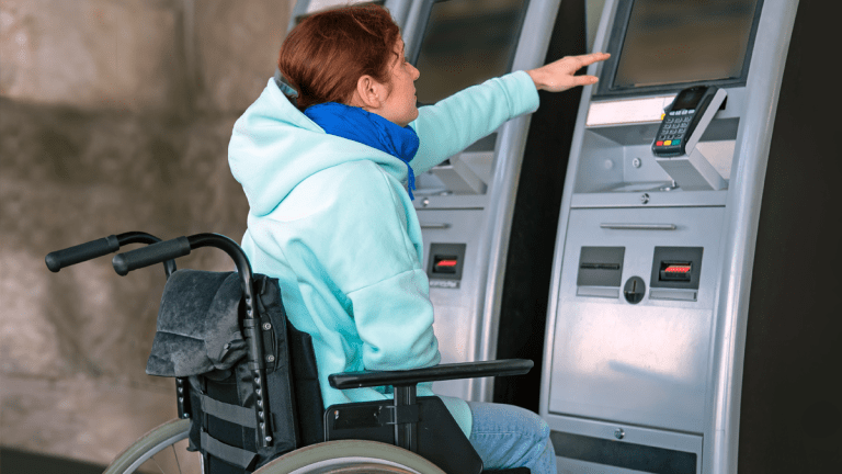 En kvinna i rullstol framför en biljettautomat. Hon försöker nå upp till skärmen, men den sitter för högt upp.
