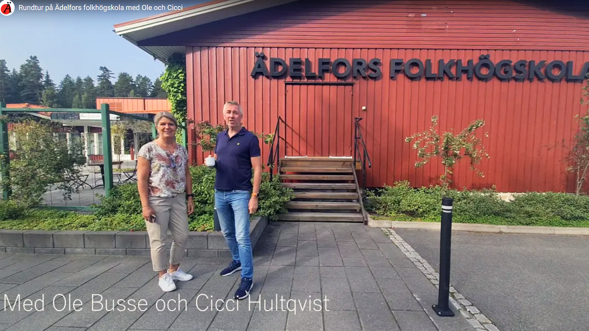 Cicci och Ole står framför en röd trävägg. På väggen finns skylten Ädelfors folkhögskola.