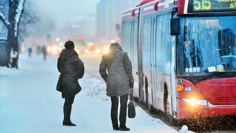 Två personer står ute i snöoväder, intill en buss, och väntar på att bli insläppta.