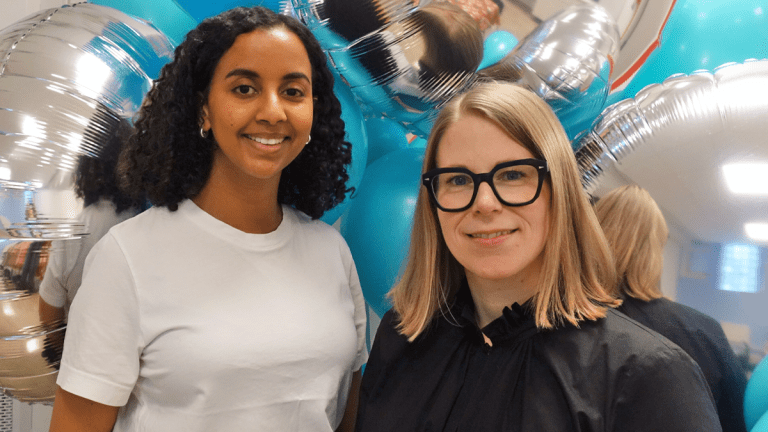 Handläggarna Salina och Karin framför en vägg av blå och silverfärgade ballonger.