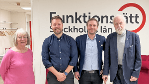 Fyra personer står på rad framför Funktionsrätt Stockholms läns logga. Till vänster en gråhårig kvinna i rosa tröja. I mitten står två yngre män klädda i blått. Till höger står ännu en man i blått, men han är lite äldre och gråhårig.