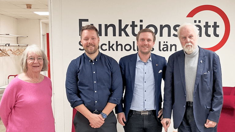 Fyra personer står på rad framför Funktionsrätt Stockholms läns logga. Till vänster en gråhårig kvinna i rosa tröja. I mitten står två yngre män klädda i blått. Till höger står ännu en man i blått, men han är lite äldre och gråhårig.