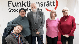 En person sitter i rullstol intill fyra personer som står på rad framför vår logga. Alla ler in i kameran.