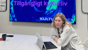 Karin Forss, en blond kvinna med vit skjorta sitter framför en dator. Bakom henne finns en skärm med en presentation.