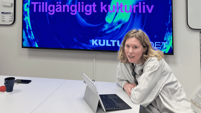 Karin Forss, en blond kvinna med vit skjorta sitter framför en dator. Bakom henne finns en skärm med en presentation.