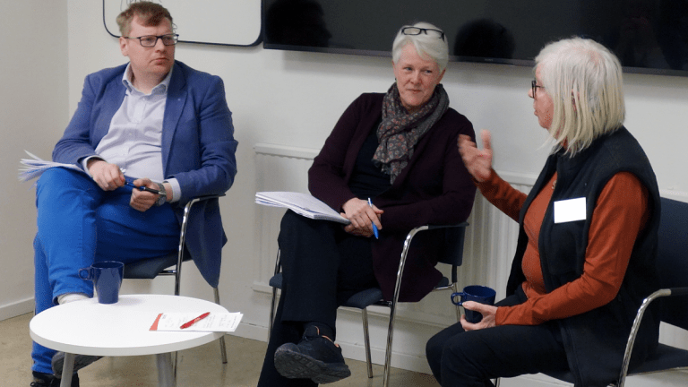 Paneldiskussion under mötet om inkludering på arbetsmarknaden. Tre personer sitter på rad längst fram i möteslokalen och diskuterar.