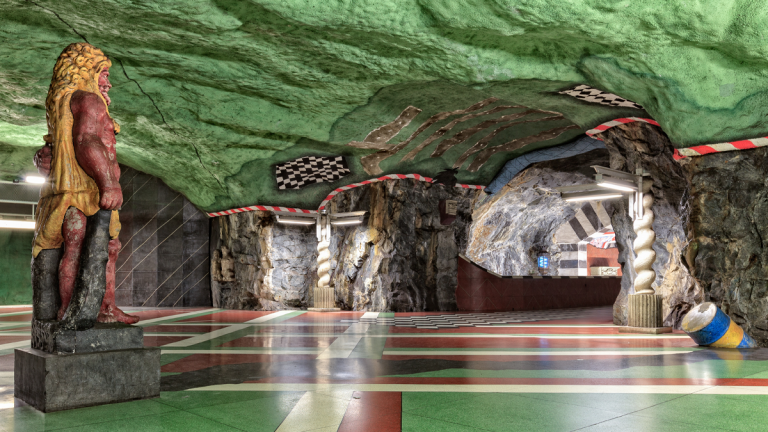 Konst på Kungsträdgårdens tunnelbanestation. En staty av en rödmålad man i gul tunika blickar mot ingången. Golvet är indelat i röda och gröna rutor och en oljetunna lutar ut från väggen. En tjock rand av mörk sten ligger som ett oljespill mellan statyn och tunnan.