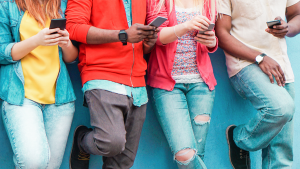 Fyra personer står på rad framför en blårappad vägg. De flesta lutar sig avslappnat mot väggen. Alla är upptagna med sina mobiltelefoner.