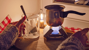 Ett tänt stearinljus lyser upp ett bord. På en bricka står en gasolbrännare. Den brinner och värmer upp en liten kastrull. Intill på brickan håller en kvinna handen om en temugg, preppad med en sked och en ny tepåse.