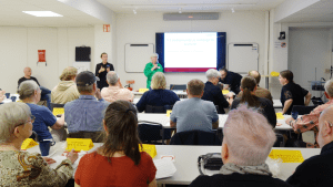 Stora rummet i ÅttiotvåAn är fullt av personer som lyssnar på Annika Hässler, som talar i en mikrofon, längst fram i salen.