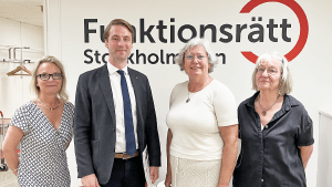 Fyra personer står på rad framför en vägg med vår logga. De ler mot kameran.