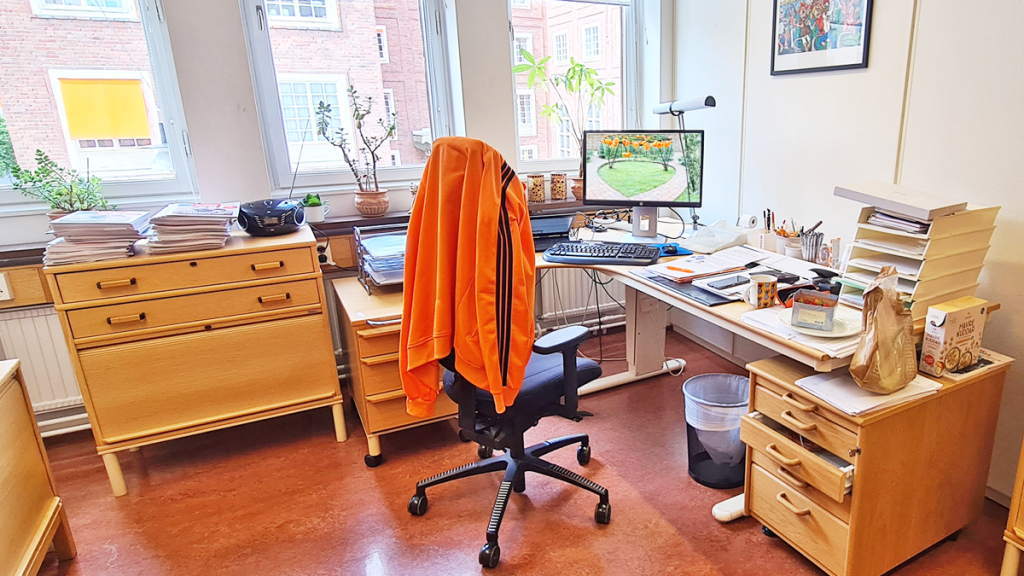 Några låga förvaringsskåp står mot en vägg med tre fönster. Skåpen bildar ett L med ett skrivbord, som står emot väggen intill till höger.