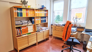 Några låga förvaringsskåp står mot en vägg med fönster. Skåpen bildar ett L med två höga fövaringsskåp, som står emot väggen intill till vänster. Längst till vänster skymtar en dörr.