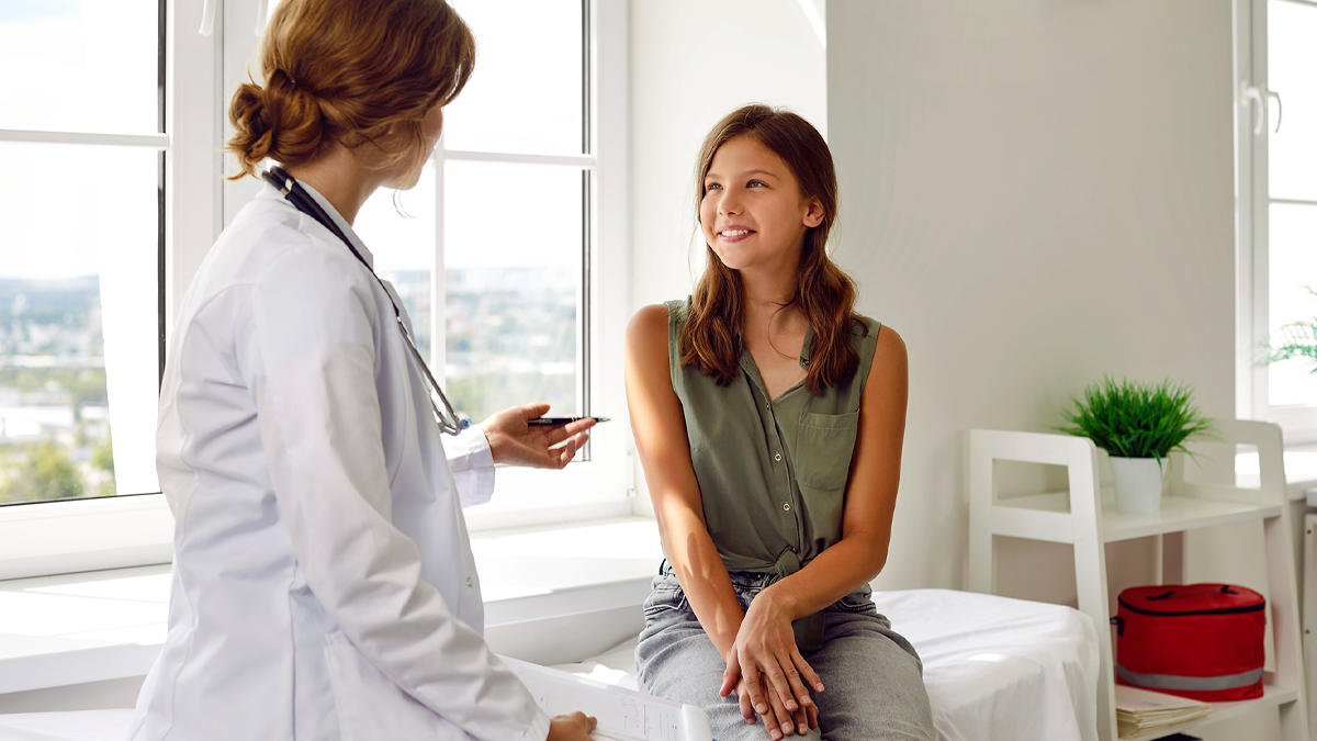 En tonåring sitter på en undersökningsbrits och ser på en läkare som står bredvid. Läkaren berättar något och tonårsflickan ler.