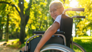 En tjej med glasögon ler mot kameran. Hon sitter i en rullstol med händerna över hjulen, redo att köra iväg. I bakgrunden solbelysta träd och ängar.