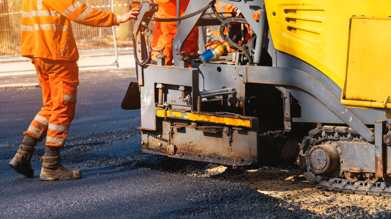 En person i orange overall står intill en maskin som lägger asfalt. Trafikverket ansvarar bland annat för vägars framkomlighet.