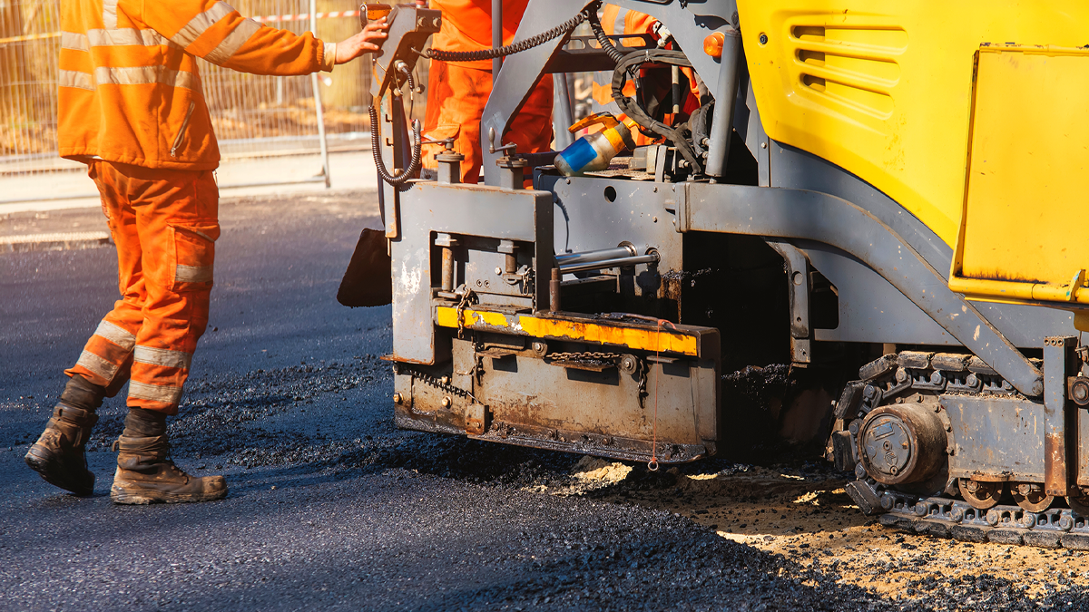 En person i orange overall står intill en maskin som lägger asfalt. Trafikverket ansvarar bland annat för vägars framkomlighet.