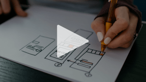 Över bilden ligger en vit spela-knapp. Bilden föreställer en hand som fyller i detaljer med en orange penna i en handritad modell av en app under utveckling.