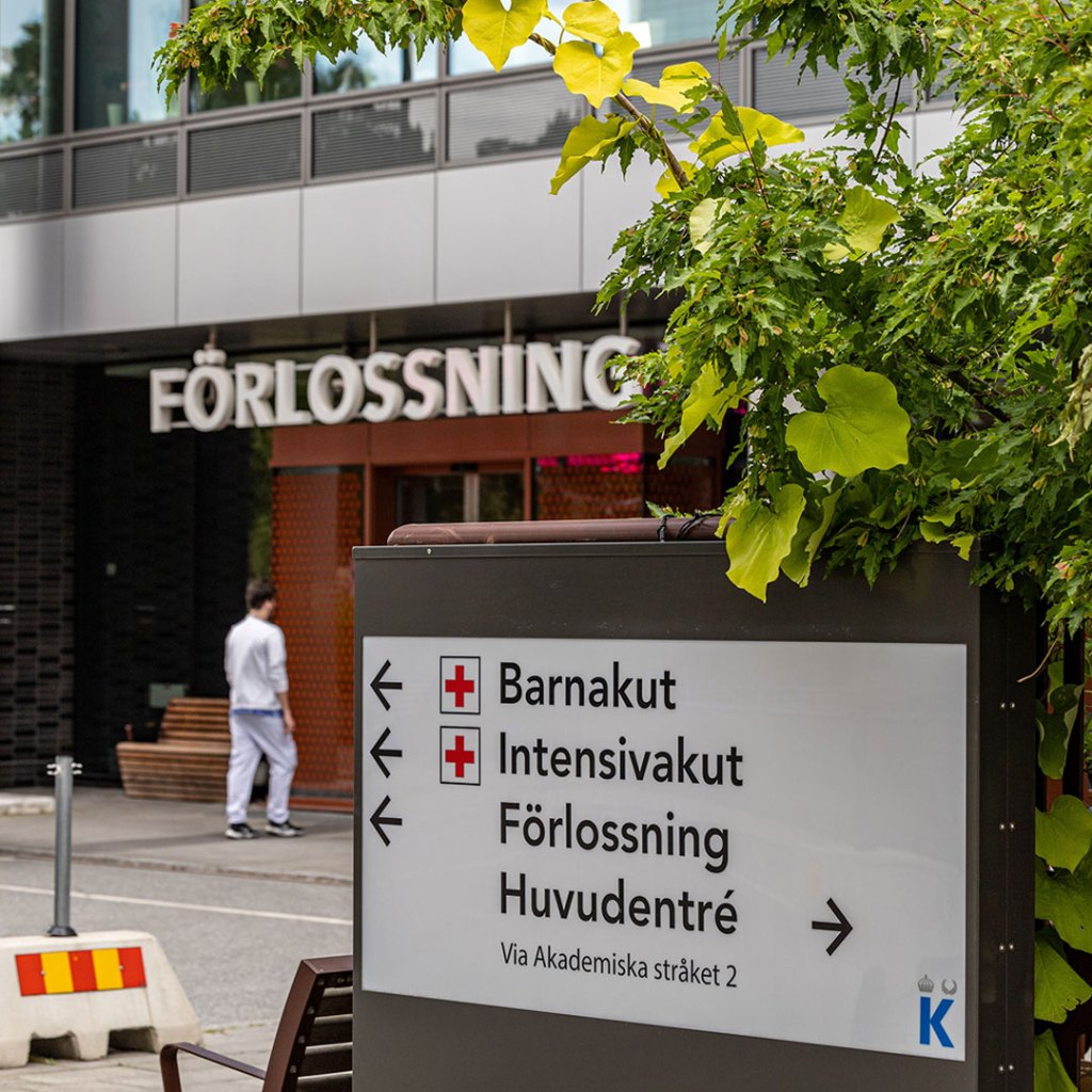 En person i vita sjukhuskläder på väg in i en entrédörr. Över dörren syns en skylt med ordet förlossning.