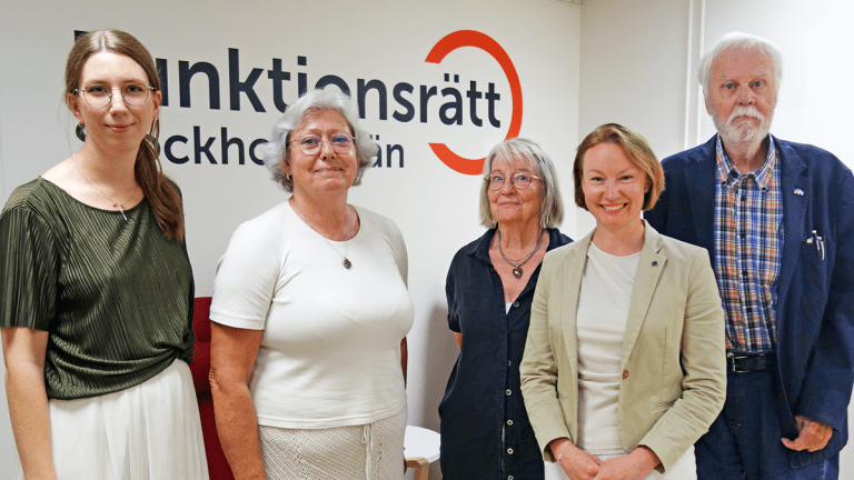 Fem personer står på rad framför en vägg med vår logga på. De ler in i kameran.