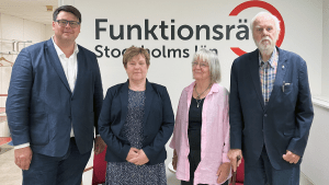 Fyra personer står på rad framför en vägg med vår logga på. De ler in i kameran.