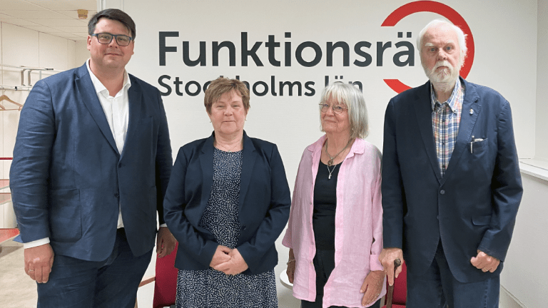 Fyra personer står på rad framför en vägg med vår logga på. De ler in i kameran.