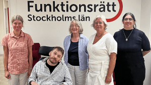 Fyra personer står på rad tillsammans med en som sitter i rullstol. Bakom dem finns en vägg med vår logga på. De ler in i kameran.