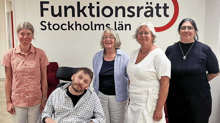 Fyra personer står på rad tillsammans med en som sitter i rullstol. Bakom dem finns en vägg med vår logga på. De ler in i kameran.