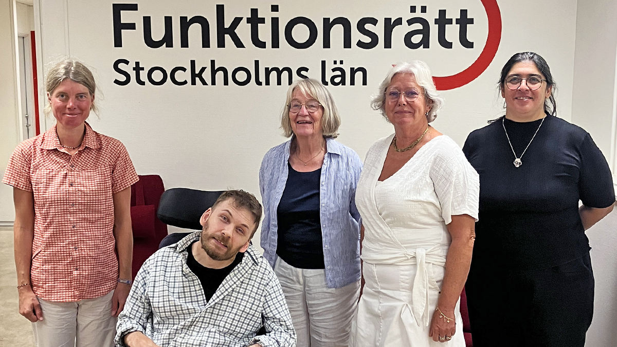 Fyra personer står på rad tillsammans med en som sitter i rullstol. Bakom dem finns en vägg med vår logga på. De ler in i kameran.