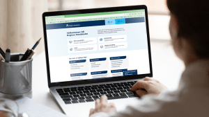 En person syns snett bakifrån. Personens händer är på tangentbordet på en laptop. På skärmen syns Region Stockholms hemsida.