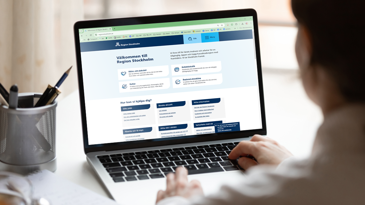 En person syns snett bakifrån. Personens händer är på tangentbordet på en laptop. På skärmen syns Region Stockholms hemsida.