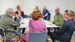 Flera personer sitter runt två ihopställda bord. En kvinna pratar och gestikulerar med händerna. Alla runt bordet tittar på henne.