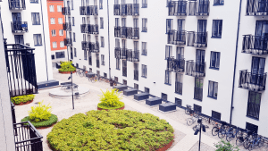 En innegård med några små runda planteringar fyllda med gröna växter står omringade av grått grus. Genom gruset skär en väg av stenplattor. Flera cyklar står parkerade i cykelställ. Runt gården reser sig vita flervåningshus, med små balkonger med svarta gallerräcken.