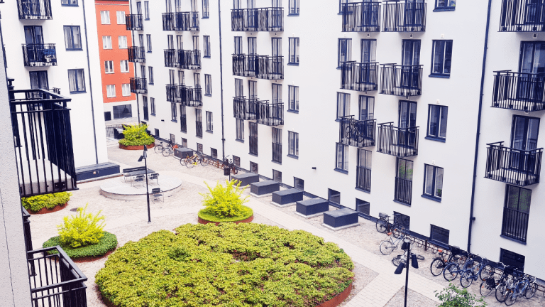En innegård med några små runda planteringar fyllda med gröna växter står omringade av grått grus. Genom gruset skär en väg av stenplattor. Flera cyklar står parkerade i cykelställ. Runt gården reser sig vita flervåningshus, med små balkonger med svarta gallerräcken.
